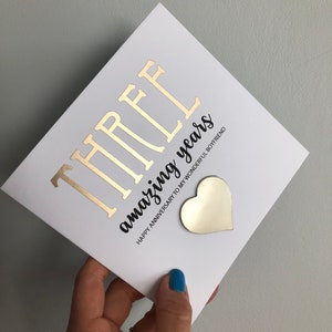 May include: A white card with gold foil lettering that reads "THREE amazing years". The card also has a gold heart sticker with the text "Happy Anniversary to my wonderful boyfriend".