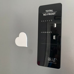 May include: A white heart-shaped magnet is attached to a stainless steel refrigerator door. The refrigerator has a digital control panel with a black background and white text that reads "TOTAL NO FROST". The control panel has a temperature display with a range of -14 to 8 degrees Celsius.