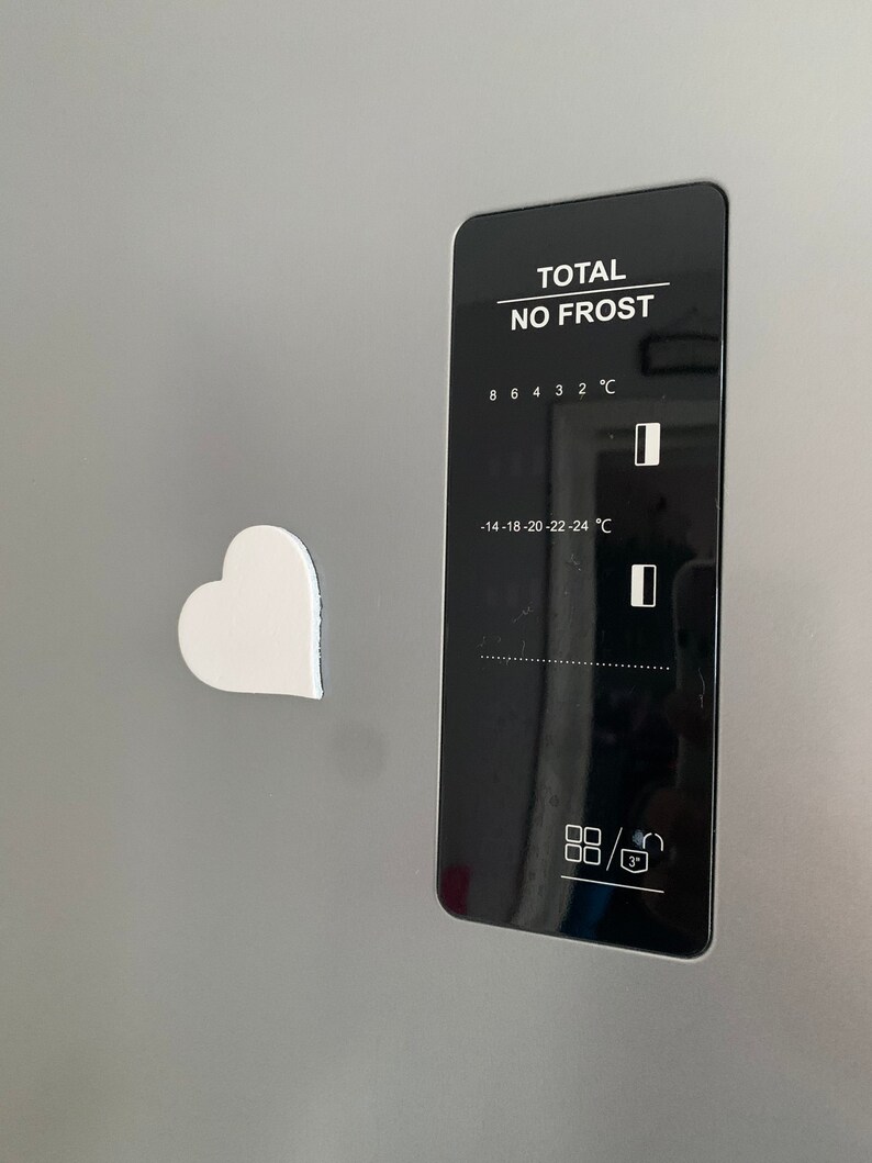 May include: A white heart-shaped magnet is attached to a stainless steel refrigerator door. The refrigerator has a digital control panel with a black background and white text that reads "TOTAL NO FROST". The control panel has a temperature display with a range of -14 to 8 degrees Celsius.