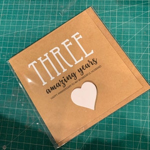May include: A brown card with the text "THREE amazing years" and "HAPPY ANNIVERSARY TO MY WONDERFUL HUSBAND". A white heart shape is cut out of the card.
