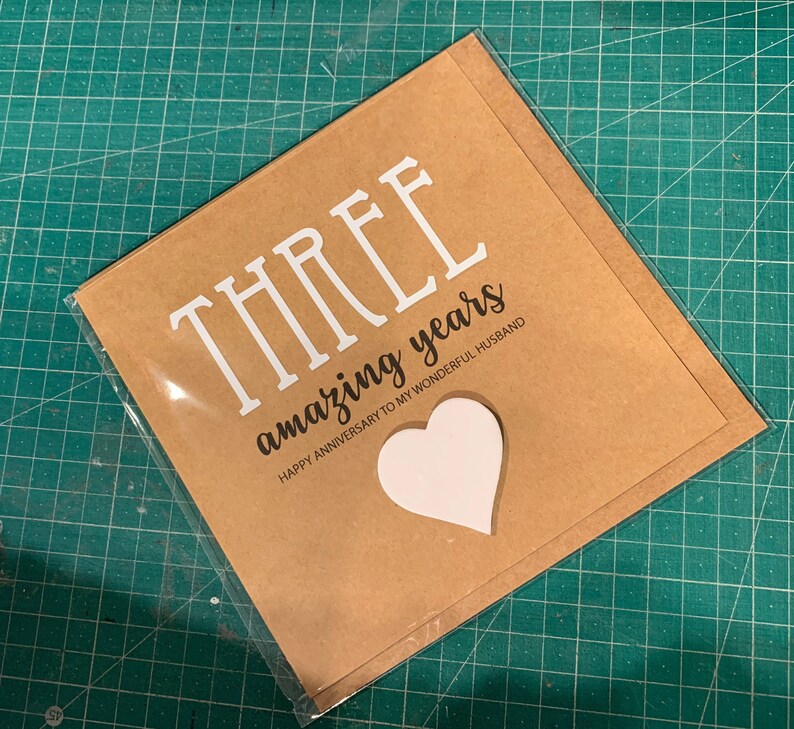 May include: A brown card with the text "THREE amazing years" and "HAPPY ANNIVERSARY TO MY WONDERFUL HUSBAND". A white heart shape is cut out of the card.