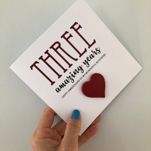 May include: A white card with the text "THREE amazing years" in black and red lettering. The card also has a red heart cutout. The text below the main text reads "Happy Anniversary to my wonderful boyfriend".