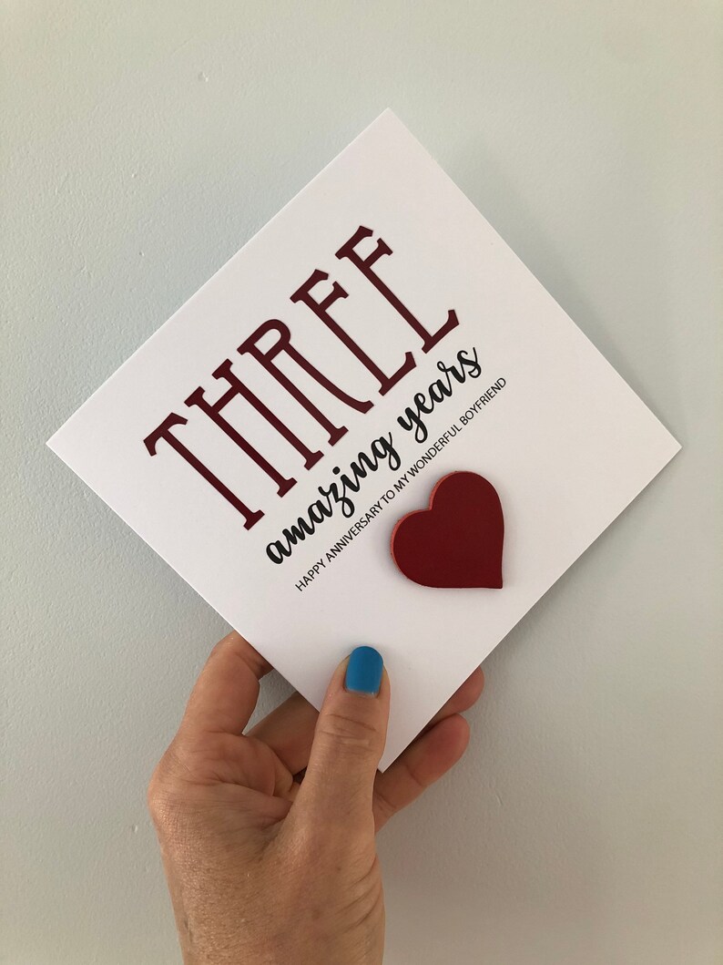 May include: A white card with the text "THREE amazing years" in black and red lettering. The card also has a red heart cutout. The text below the main text reads "Happy Anniversary to my wonderful boyfriend".