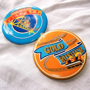May include: Two round, vintage pinback buttons. One is blue with "Magic Mountain Freefall" text and a roller coaster graphic. The other is orange with "Gold Rusher" text and a roller coaster car graphic.