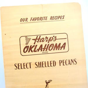 May include: A light tan rectangular paper label with dark brown text reading OUR FAVORITE RECIPES, Harp's OKLAHOMA BRAND, and SELECT SHELLED PECANS. The logo features a small harp icon, and a small illustration of a person holding a tray is at the bottom.