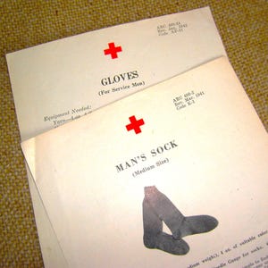 May include: Two vintage American Red Cross instructional pamphlets with off-white paper. The top document is titled GLOVES (For Service Men) with a red cross. The bottom document is titled MAN'S SOCK (Medium Size) with a red cross and a sock illustration.
