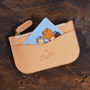 May include: A light tan leather cardholder with a zipper closure and a card peeking out. The card has a cartoon illustration of a bear and a man. The cardholder has the logo "Ranta" printed on it.