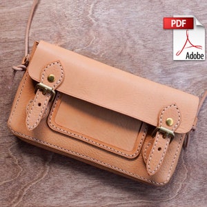 May include: A light tan leather crossbody bag with a rectangular shape. The bag features a front flap with two brass buckles and a small front pocket. The stitching is visible in a lighter colour, and the bag has a long, thin strap.