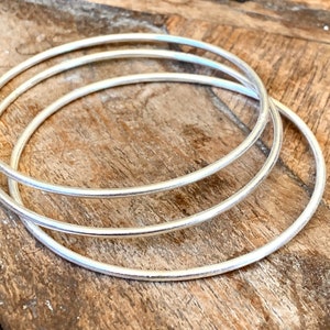 May include: Three silver bangles are stacked on a rustic wooden surface. The thin, circular bracelets are made of a shiny, metallic material. The bangles are simple in design and appear to be lightweight.