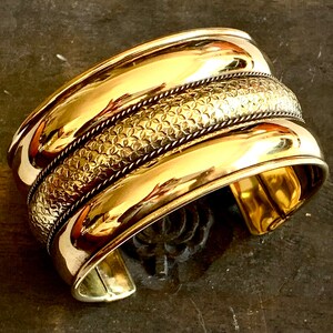 May include: A wide gold-toned metal cuff bracelet with a textured band and a smooth, polished finish.