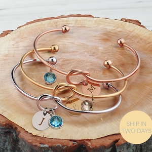 May include: Three gold, rose gold, and silver knot bangle bracelets with initial charms and blue crystal accents. The bracelets are stacked on a wooden surface.