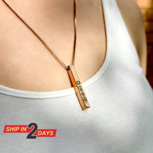 May include: A gold chain necklace with a vertical bar pendant. The pendant has a small circle at the top and a series of lines engraved on the front.