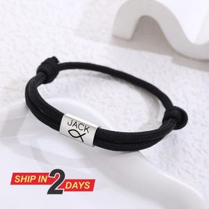May include: Black braided bracelet featuring a silver-coloured cylindrical bead. The bead is engraved with the name "JACK" and a fish symbol. The bracelet is knotted at both ends.