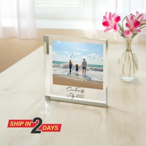 Peut inclure: Un bloc photo en acrylique transparent affichant une photo de famille sur une plage. La photo montre une famille de trois personnes debout dans l'océan. Le texte "Our Family July 2025" est imprimé sur la photo. Un vase de fleurs roses est en arrière-plan.