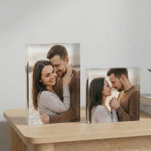 May include: Two clear acrylic photo blocks displaying a couple embracing. The larger block shows the couple hugging, while the smaller block shows them face to face. The photos are set on a light-coloured wooden surface.