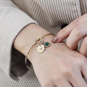 May include: Gold bangle bracelet with a knotted charm and a small round charm engraved with the letter "M". A green gemstone is attached to the knotted charm.