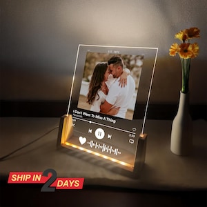 May include: A clear acrylic photo display with a wooden base, illuminated by LED lights. The display features a photo of a couple and a music player interface with the song title "I Don't Want To Miss A Thing". The text "EST. 2023" is also visible.