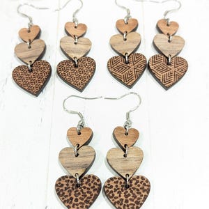 May include: Wooden heart earrings with silver hooks. The earrings feature three heart shapes in varying shades of brown. Some hearts have geometric patterns, while others have a leopard print design. The earrings are lightweight and suitable for everyday wear.