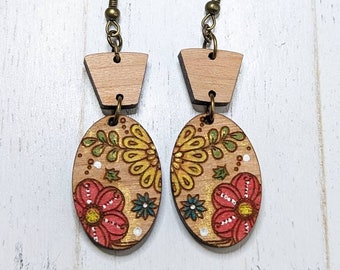 May include: A pair of wooden earrings with a floral design. The earrings are oval shaped and have a small, rectangular piece of wood at the top. The flowers are painted in shades of red, yellow, and green.