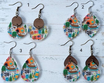 May include: Four pairs of camping themed earrings. The earrings are made of wood and acrylic. The acrylic pieces feature a camping scene with a camper, trees, and a lake. The earrings are available in four different styles.