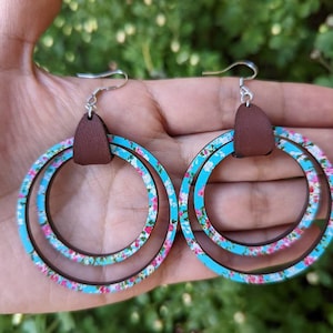 May include: A pair of blue and white floral patterned wooden hoop earrings with brown leather accents. The earrings have a double hoop design.