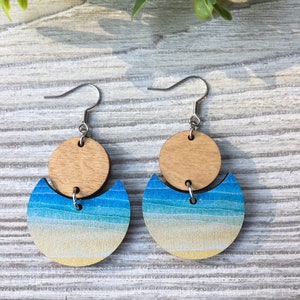 May include: A pair of wooden earrings with a beach scene design. The earrings are made of two circles, one on top of the other. The top circle is a natural wood color, and the bottom circle is painted with a gradient of blue, green, and yellow to represent the ocean and sand. The earrings are attached to silver hooks.