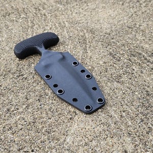 May include: A black push dagger with a textured handle and a sheath. The dagger has a T-shaped handle and a double-edged blade. The sheath has multiple eyelets for attachment. The items are on a textured, gray surface.