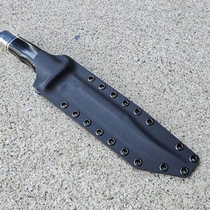 Puede incluir: Una funda de cuchillo de cuero negro con una punta de metal plateado y costuras negras. La funda tiene múltiples ojales a lo largo del borde.