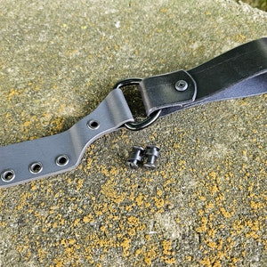 May include: A grey leather belt loop with four metal eyelets and a black leather strap with a metal D-ring. Two black metal rivets are also visible.