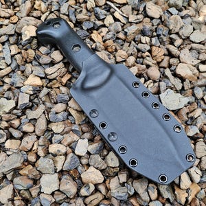 Könnte beinhalten: Eine schwarze, geformte Messerscheide für ein Messer mit feststehender Klinge, mit einem konturierten Griffbereich und einem breiten, flachen Klingengehäuse mit einer Reihe von Metallösen an den Kanten, gefertigt aus robustem, mattem Kunststoff.