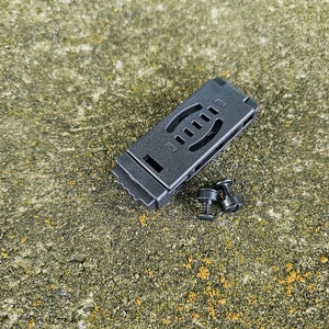 Pode incluir: Clip de plástico preto com dois parafusos pretos. O clip tem uma superfície texturizada e uma série de furos.