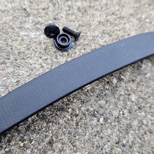Op de afbeelding: Een gebogen zwarte leren riem met een knoop en een gat, samen met kleine zwarte schroeven en bevestigingsmiddelen. De items liggen op een gestructureerd, grijs oppervlak. De riem is waarschijnlijk voor een tas.