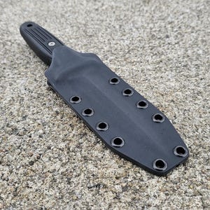 Puede incluir: Un cuchillo negro con una funda negra. El cuchillo tiene un mango texturizado y una hoja puntiaguda. La funda tiene múltiples ojales para la fijación. El cuchillo y la funda están sobre una superficie gris texturizada.