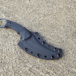 May include: A black karambit knife with a finger ring and a black sheath. The knife is made of metal and has a curved blade. The sheath has multiple eyelets for attachment.