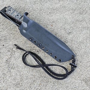 May include: A black knife sheath with a brown leather strap and a black cord. The sheath has a textured surface and is made of a durable material.
