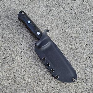 May include: A black knife with a textured handle and two white dot accents, sheathed in a black holder. The knife and sheath are set against a textured gray surface.