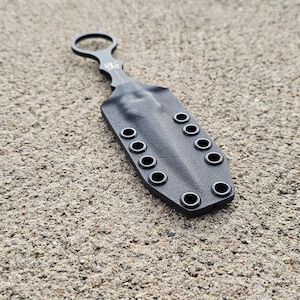 May include: A black, small knife with a ring handle and a sheath. The sheath has multiple eyelets along the sides. The knife and sheath are lying on a textured, light brown surface.