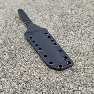 Puede incluir: Una funda de cuchillo negra con múltiples ojales para sujeción. La funda está hecha de un material duradero y tiene un mango texturizado. El diseño es simple y funcional, adecuado para uso en exteriores o táctico.