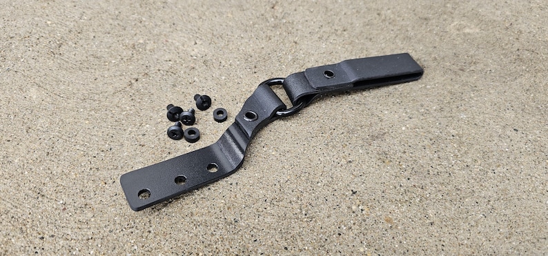 May include: Black metal belt clip with a curved design and multiple holes. The clip has a small loop at the top and comes with several black screws.