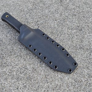 Puede incluir: Una funda de cuchillo de cuero negro con múltiples filas de costuras y un cierre de presión.