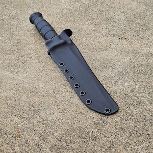 Könnte beinhalten: Ein schwarzes Messer mit strukturiertem Griff und passender Scheide. Die Scheide hat mehrere Ösen an der Seite. Das Messer und die Scheide liegen auf einer rauen, hellbraunen Oberfläche.