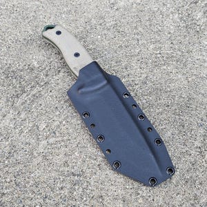 Puede incluir: Una funda de cuchillo de cuero negro con múltiples filas de costuras y un cierre a presión. La funda está diseñada para sujetar un cuchillo de hoja fija.