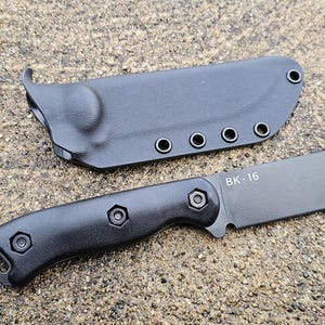 May include: A black knife and sheath set. The knife features a dark blade and a black handle with hexagonal screws. The sheath is also black and has several eyelets. The blade is marked with the text "BK-16".