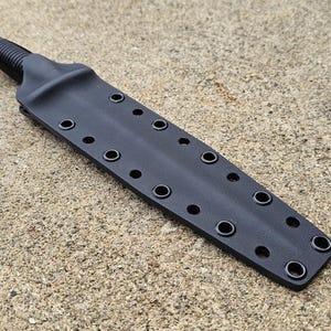 Puede incluir: Un cuchillo negro con un mango texturizado y una funda. La funda tiene múltiples ojales a lo largo de los bordes. El cuchillo y la funda están sobre una superficie texturizada de color claro.