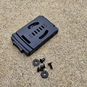 Pode incluir: Uma coldre de plástico preto com várias seções recortadas e uma série de pequenos parafusos e arruelas. O coldre é retangular com uma superfície texturizada e foi projetado para segurar um item. O hardware também é preto.