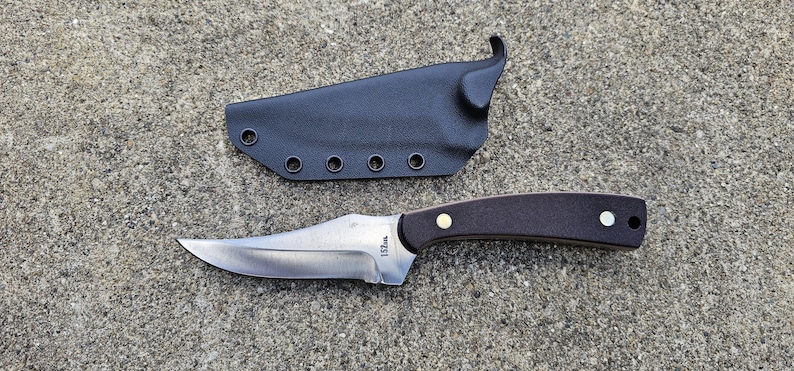 May include: A black leather knife sheath with a brown wooden handle knife with a silver blade. The blade has the text "152mm" on it.