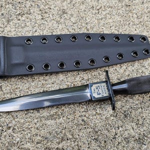May include: A black Fairbairn-Sykes fighting knife with a double-edged blade and a textured handle. The blade features a small rectangular plate engraved with the text The F-S Fighting Knife. A black sheath with metal eyelets is included.