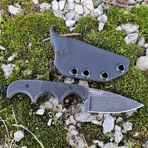 Può includere: Un coltello nero a lama fissa con fodero abbinato, appoggiato su un letto di muschio verde e piccole rocce. Il coltello ha un'impugnatura strutturata e un cordino. Il fodero ha più occhielli.