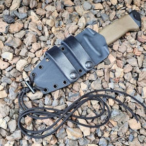 Puede incluir: Una funda de cuchillo de cuero negro con un cuchillo de mango marrón en su interior. La funda tiene una correa y un lazo para sujetarla a un cinturón.