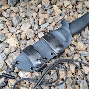 May include: A black knife with a black handle and a black sheath. The sheath has two straps with snap closures. A black cord is attached to the sheath. The knife and sheath are resting on a bed of small rocks.
